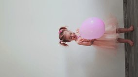 Adorable two years old birthday girl in dress deflating balloon. Home birthday celebration, minimalist style. Vertical video