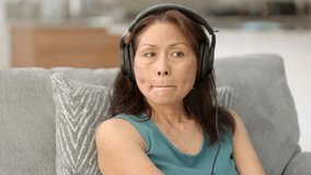 Close up attractive woman wearing headphones enjoying listening to music moving head to song looking at television sitting on comfortable sofa at home. Mature asian female watching music video.