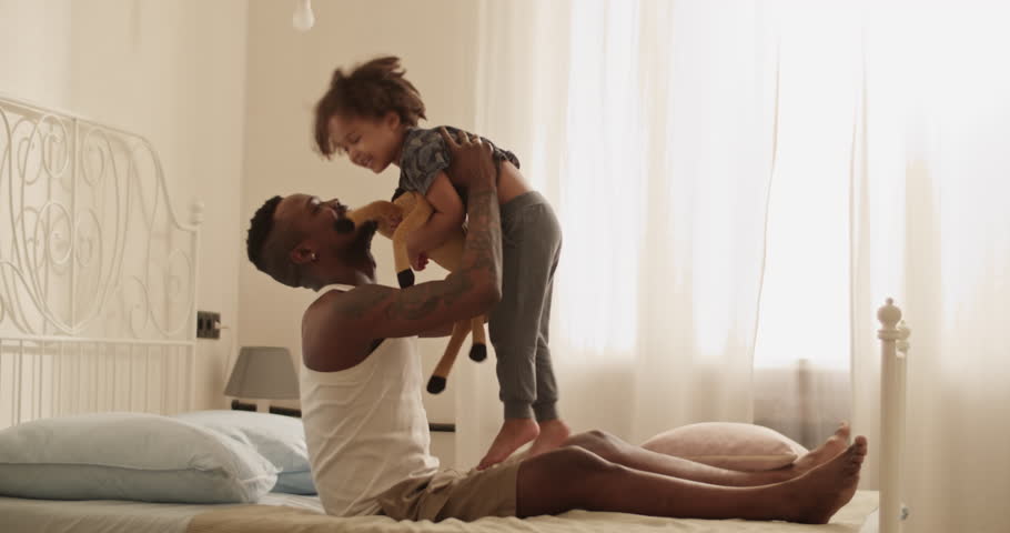 Delighted African American man playing with cute daughter on bed