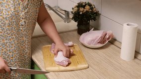 Video of cropped woman in bathrobe cutting, splitting chicken meat flesh with knife and putting legs in white plate on wooden drawer in kitchen. Household chores, cooking and preparation of lunch