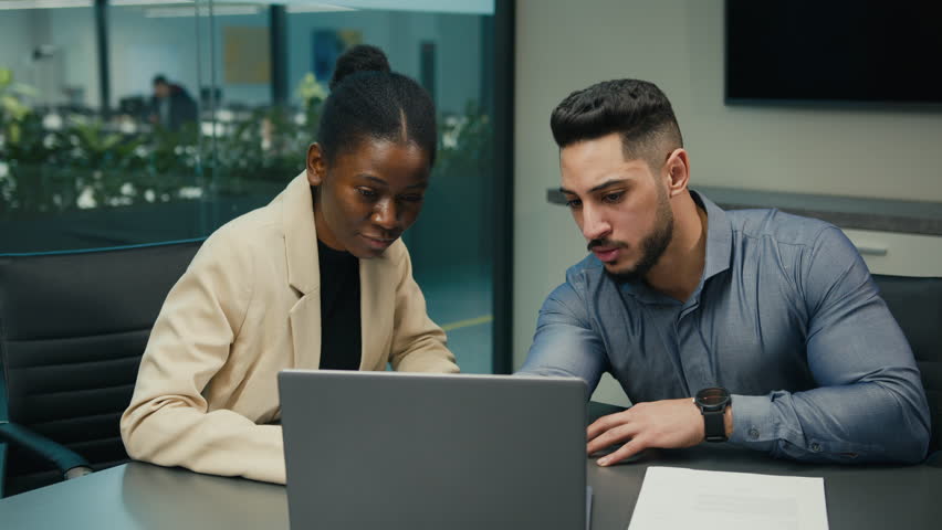Diverse Indian Arabian man and African American woman colleagues discuss financial data analyzing documents marketing project result in report use laptop talk business conversation sit at office desk Royalty-Free Stock Footage #1102429665