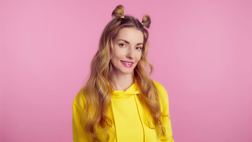 Woman sends air kiss smiling looking at camera. Happy positive emotional blonde girl. Joyful cheerful pretty lady in yellow hoodie, smile face. Indoors studio, isolated on pink background | Shutterstock HD Video #1102434019