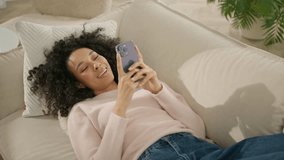 Smiling African American woman holding smart phone at home in sunlight. Woman of color watching social media lying on comfortable sofa. Portrait of curly girl, smartphone entertainment in slow motion