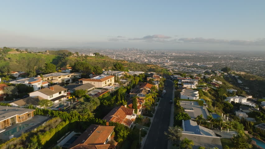 Forwards fly above deluxe homes in urban area situated on hills above metropolis lit by golden hour sunrays. Los Angeles, California, USA