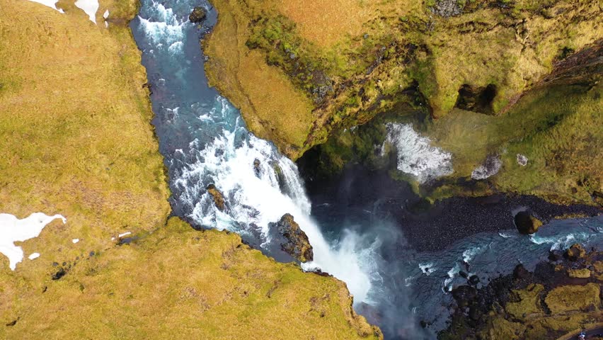 aerial view from kvernufoss waterfall. the drone flies from the meadow over the cliffs where the waterfall flows down. the waterfall is surrounded by a green-yellow meadow. water shoots down the gorge
