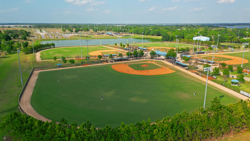 Two Baseball Teams Playing Game On The Green Field Of Lake Myrtle Sports Park In Auburndale, Florida Under Blue Skies. High Angle Drone Orbiting Shot