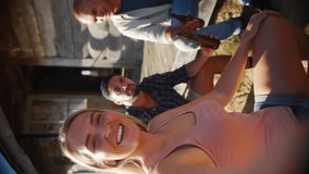 Vertical video POV shot of group of friends sitting on porch of cabin in countryside drinking beer and posing for selfie - shot in slow motion