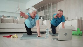 Online training of senior couple at home with laptop. Aged family man and woman doing physical exercises on mats in living room. Smiling elderly people training, having yoga pilates practice together.