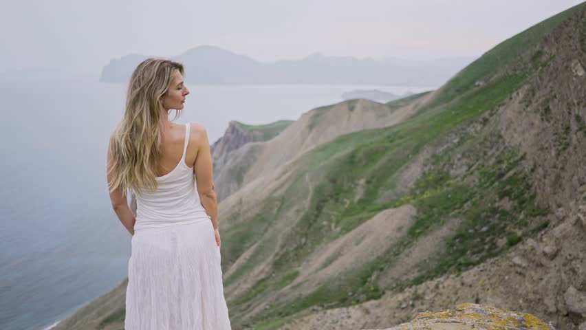Young woman blonde in white elegant dress standing on rocky cliff edge enjoying tranquil and calmness against mountains and blurred sea close backside view