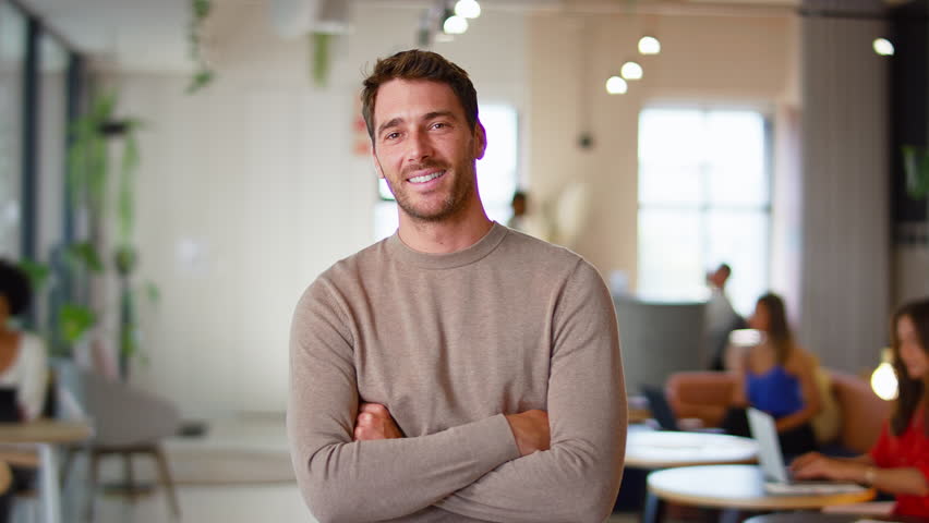 Portrait of smiling businessman standing in modern open plan office looking into camera with folded arms - shot in slow motion