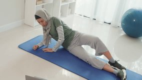 Sporty youth Asia islam woman wear muslim sportswear exercise stretch her legs and flow arm beginner practice online class training from laptop at home in living room. healthcare and fitness activity.