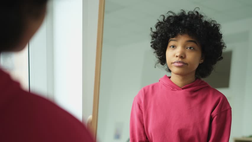 Love yourself. Beautiful young smiling african american woman dancing touching curly hair enjoying her mirror reflection. Black lady looking at mirror looking confident and happy. Self love concept