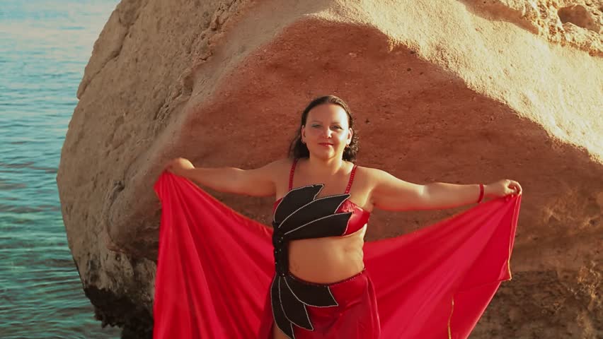 A young brunette in a red suit dances against the backdrop of the sea oriental dance playing with a large headscarf. Overall plan