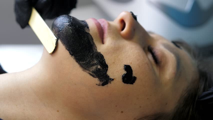 Beautician applies black mask on face of beautiful woman. Girl in spa salon doing carbon peeling procedure. Carbon peeling of the face. Skin care cosmetology. Laser therapy and skin treatment