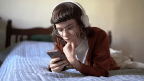 Smiling teen hipster girl wearing glasses using smartphone while relaxing at home, lying on bed scrolling social media on mobile phone, happy millennial woman in headphones listening podcast online - Powered by Shutterstock - Get 15% off with code: PIKWIZARD15