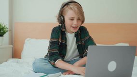 Teenage child boy using laptop and headphones. Child looking online video course lesson in bed. Chat video conference Schoolboy studying doing homework. E-learning, remote educational classes