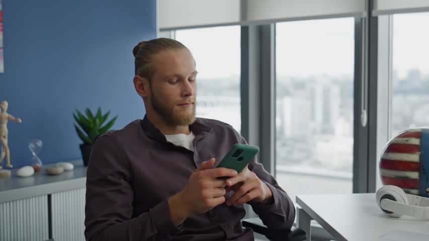 Smiling student typing mobile at table close up. Positive freelancer reading email resting smartphone at blue wall apartment. Calm man writing cellphone message taking break at big windows workplace