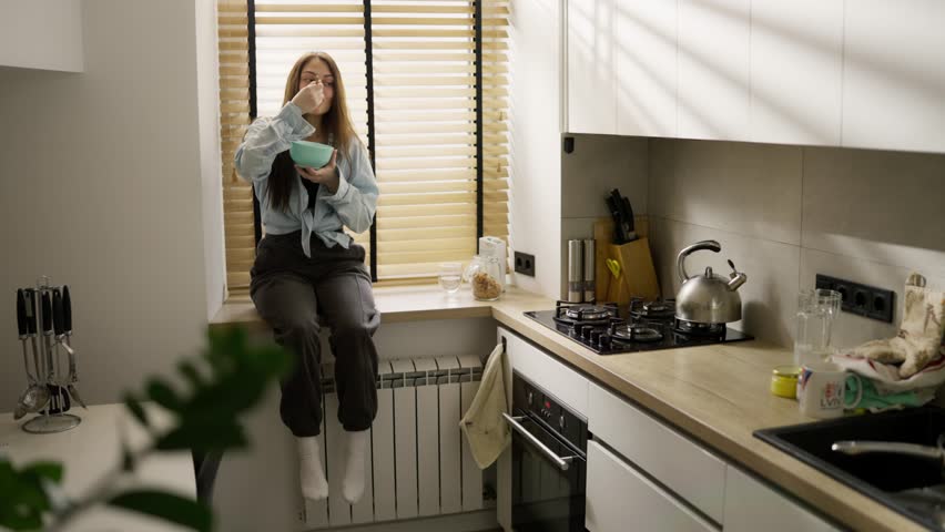 Young carefree woman eats cereal bowl sitting on windowsill at home kitchen, slow motion