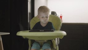 Little boy chooses video on tablet and swings legs in comfortable high chair. Surprised child opens online game app on digital device at home
