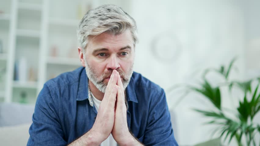 Close up portrait. Thoughtful mature gray haired bearded man sitting on the couch in the living room at home. Male in casual clothes is thinking about a problem with his hands folded and looking away Royalty-Free Stock Footage #1102507093
