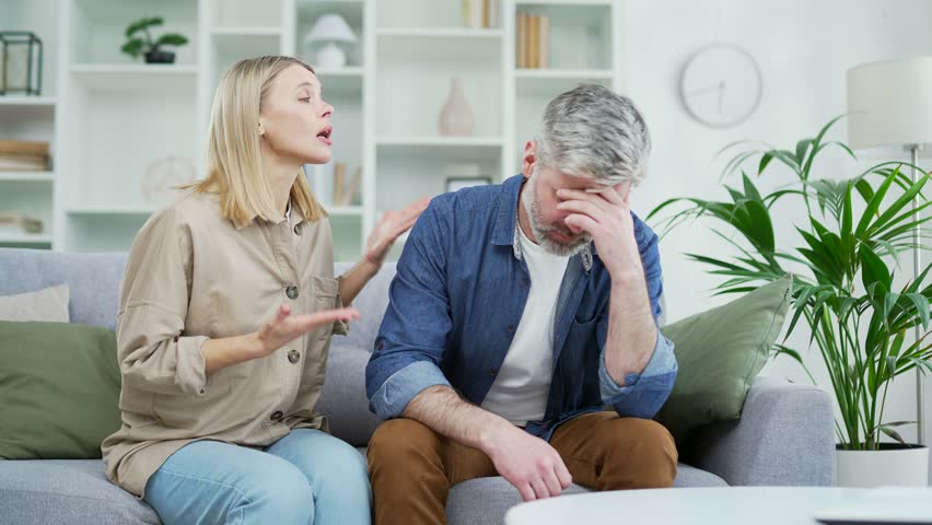 Angry wife arguing with mature husband sitting on sofa in living room at home. Conflict and dispute between a married couple. A woman quarrels with a sad man. Problem and crisis in family relations Royalty-Free Stock Footage #1102507135