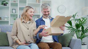 An excited married couple of a customer opens a package at home. Happy husband and wife buyers unpacking purchase sitting on sofa. Satisfied clients of e-commerce. Express delivery services concept - Powered by Shutterstock - Get 15% off with code: PIKWIZARD15