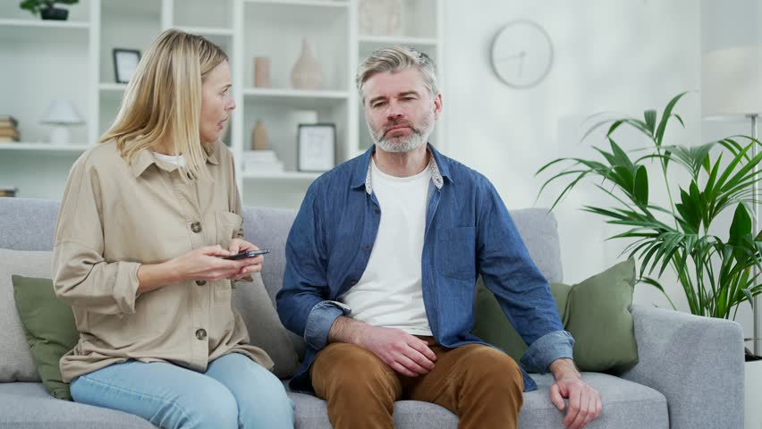 Mature gray haired man having a heart attack while sitting on the sofa in the room at home. The wife supports her husband and calls an ambulance by phone. The male is breathing hard, holding his chest Royalty-Free Stock Footage #1102507145