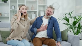 Mature gray haired man having a heart attack while sitting on the sofa in the room at home. The wife supports her husband and calls an ambulance by phone. The male is breathing hard, holding his chest - Powered by Shutterstock - Get 15% off with code: PIKWIZARD15