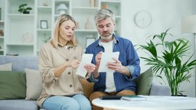 Unhappy shocked married couple looking at large utility bills sitting on sofa at home. Mature sad wife holds a payment check in her hands, and her husband writes down the data in a notebook with a pen - Powered by Shutterstock - Get 15% off with code: PIKWIZARD15