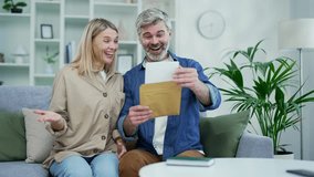 Happy married couple received a letter with good news while sitting on the couch at home. Mature smiling husband and wife open letter and read nice notification, smile, celebrate, show triumph gesture - Powered by Shutterstock - Get 15% off with code: PIKWIZARD15