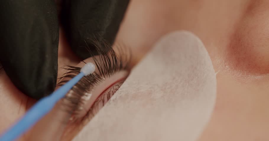 The master applies special glue before the eyelash curling procedure in pink rubber gloves close up. Master combing woman's eyelashes after lamination. Beautiful young woman with eyelash extension. Royalty-Free Stock Footage #1102521515