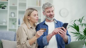 Happy married couple doing online shopping by entering credit card number on smartphone while sitting on sofa at home. A mature smiling wife and husband are satisfied with a good promotion on goods - Powered by Shutterstock - Get 15% off with code: PIKWIZARD15