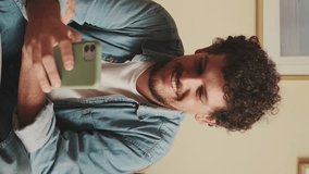 Vertical video, Happy guy browsing photos, videos on mobile phone while sitting on sofa in living room