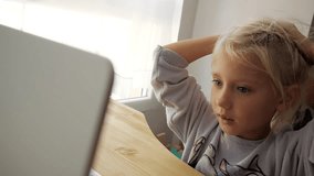 Blond caucasian child studying online. Elementary school girl grimaces at laptop's screen. Close-up