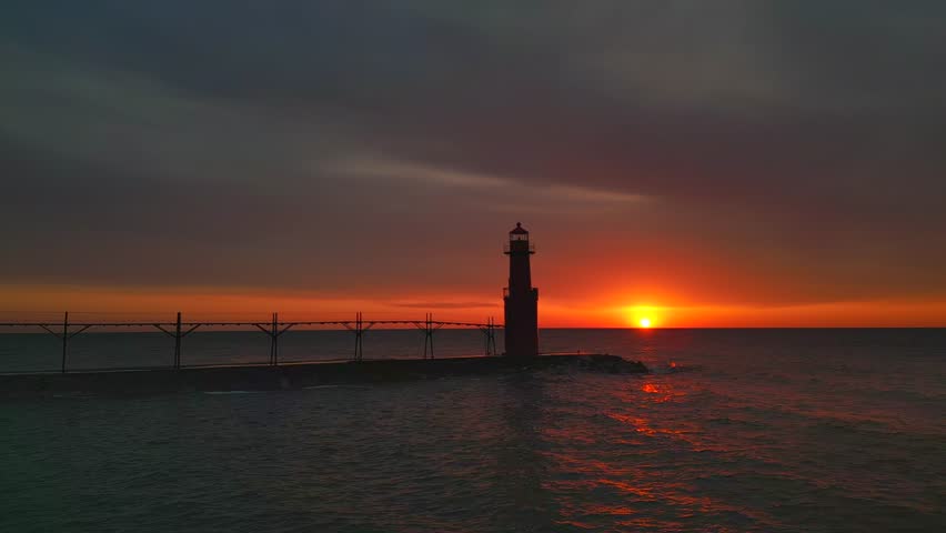 Iconic Lighthouse beacon silhouetted against fiery Lake Michigan twilight horizon, moving aerial view.