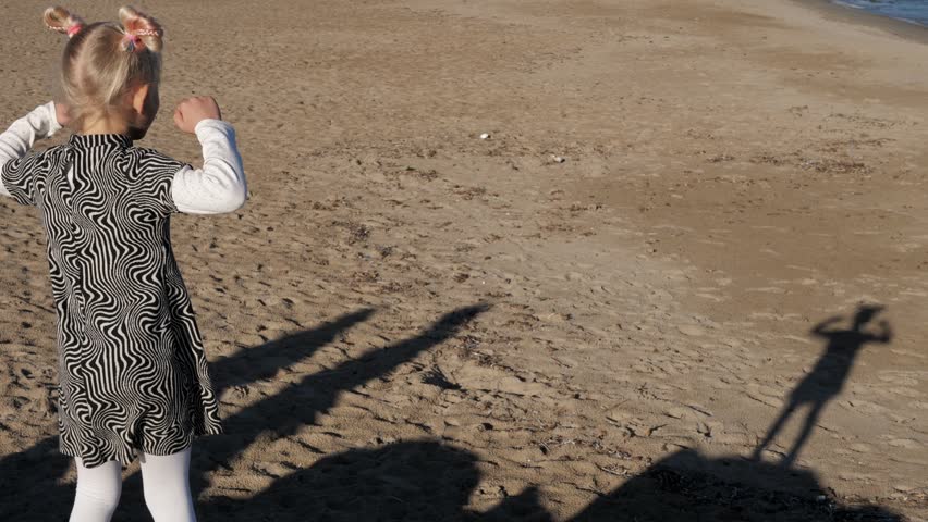 Blonde girl with double pigtails buns dances on the beach looking on her dancing shadow on the sand. Medium shot