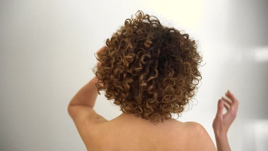Rear view of female model, with curly afro hair, looking at the camera while playing with her hands on her head. Caucasian woman with bare shoulders Royalty-Free Stock Footage #1102525653