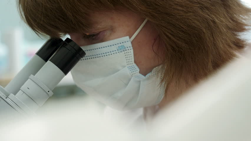 Woman looks through microscope in laboratory. Stock footage. Masked woman looks at biochemical substance through microscope. Analysis of substance under microscope in laboratory Royalty-Free Stock Footage #1102526069