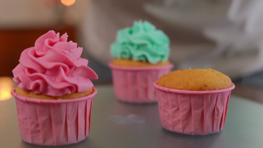 Closeup muffins decorating with pink butter cream frosting indoors. Unrecognizable confectioner pouring delicious topping on tasty pastry close-up