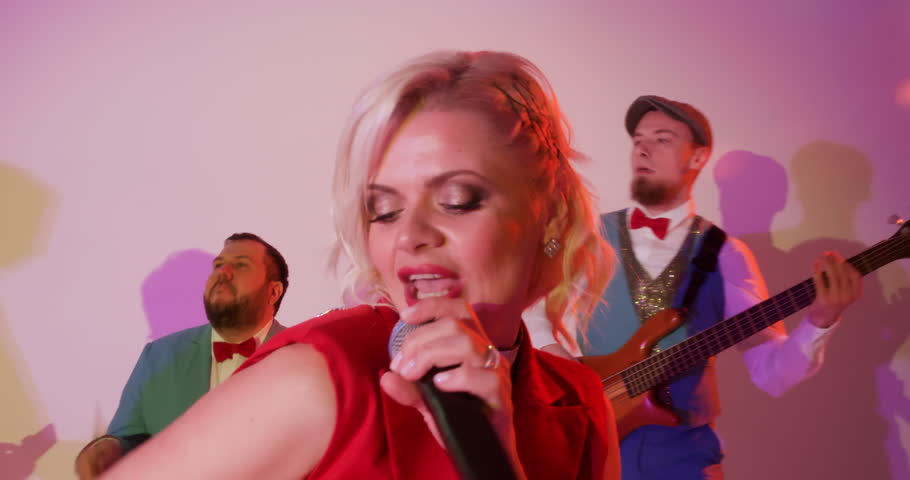 Sensual blonde woman singer in red suit emotionally sings song on stage in microphone with dancing band of musicians. Portrait of charismatic vocalist with red lips performs song in live concert. 