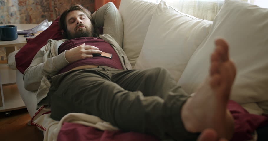 Carefree, careless lazy beard man lying on the couch napping, falling asleep, watching smartphone. Unkept, shabby look. Waste time and motivation problem. Laziness and procrastination concept Royalty-Free Stock Footage #1102529989