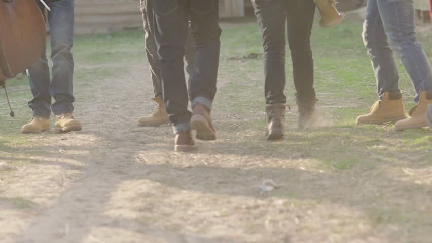 Legs of several men in jeans and brown boots walking on a dirt road kicking up dust. Slow motion. Close-up.