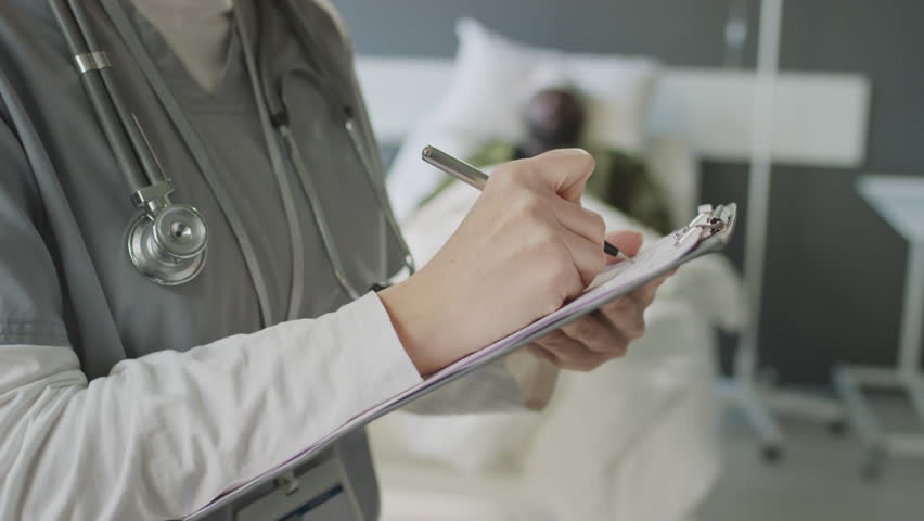 Tilt up medium close-up of Caucasian female practitioner standing military hospital room filling in patients medical card