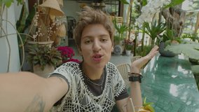 POV of young cheerful woman holding phone in outstretched arm and talking on camera while filming vlog or streaming in outdoor cafe on summer day