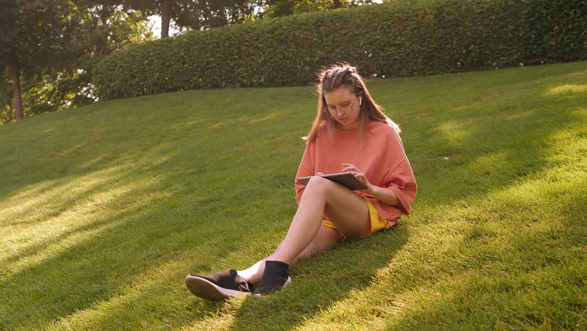 A teenage girl is sitting in a modern crowded park on the green grass with a tablet.