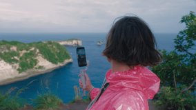 A blogger Asian woman with short trendy hair and stabilizer with phone in hand standing on edge of cliff above blue ocean and smiling on a camera. Travel trip for happy female on seaside. View point. 