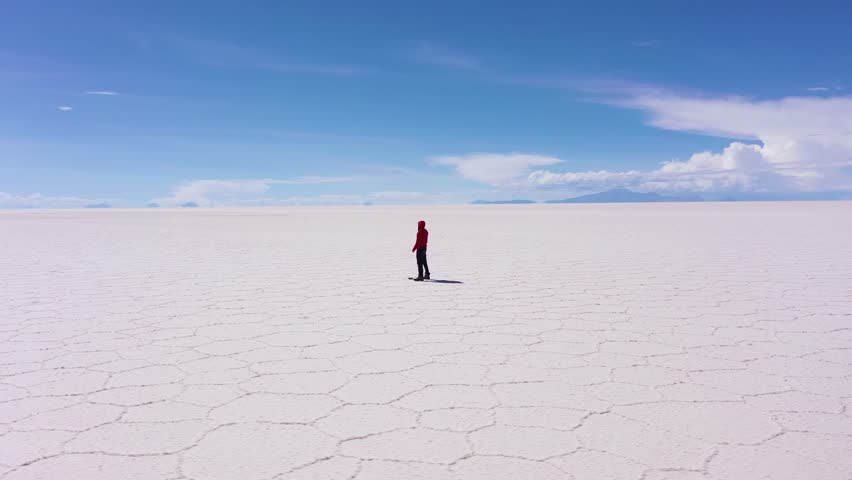 Traveler Man Looking at Uyuni Salt Flats on Sunny Day. Aerial View. Altiplano, Bolivia. Dry Season. Hexagonal Salt Formations, Cracks and Patterns. Orbiting. Wide Shot. Solo Travelling Concept