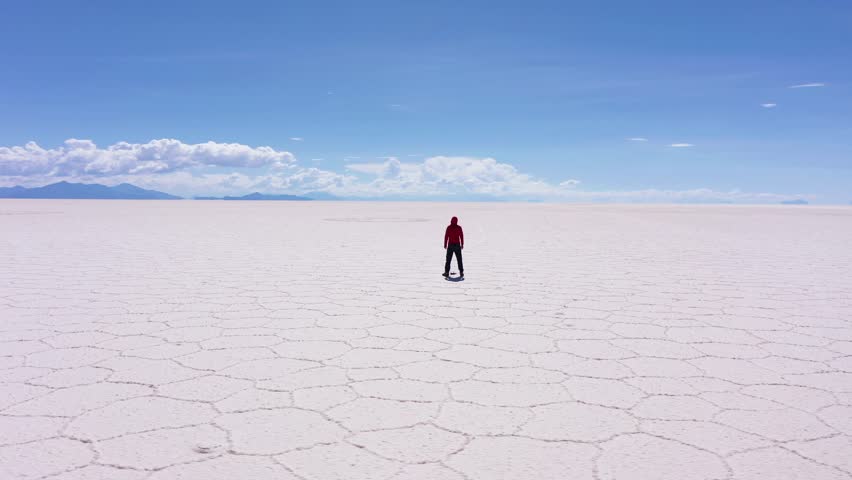 Traveler Man Looking at Uyuni Salt Flats on Sunny Day. Aerial View. Altiplano, Bolivia. Dry Season. Hexagonal Salt Formations, Cracks and Patterns. Orbiting. Wide Shot. Solo Travelling Concept