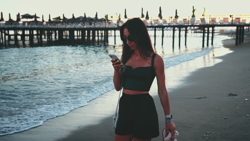 Woman with phone walks barefoot along shore beach and walks along sea. Travel weekend on landscape outdoor nature.
