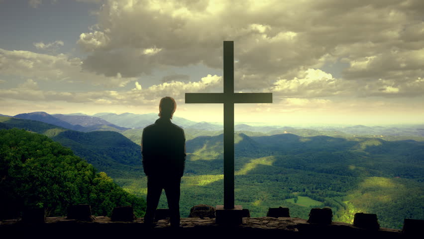 Cross Mountains Man View Cloudy Sky Flat Land Zoom In. Man standing next to a cross on top of a tower over the mountains under a cloudy sky.